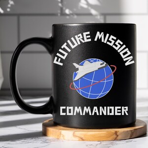 May include: Black ceramic mug with a white graphic of a space shuttle orbiting a blue globe. The text "Future Mission Commander" is printed in white on the mug.