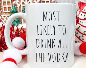 May include: White ceramic coffee mug with black text that reads "Most Likely To Drink All The Vodka". The mug is sitting on a white surface with red and white decorations.