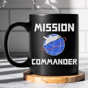 May include: Black ceramic mug with a white design that says "Mission Commander" with a space shuttle orbiting a blue globe.