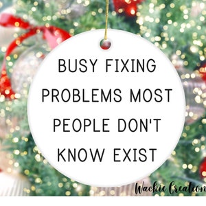 May include: White ceramic ornament with black text that reads "Busy fixing problems most people don't know exist".