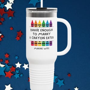 May include: White travel mug with a handle and a lid, featuring the text "BRAVE ENOUGH TO MARRY A CRAYON EATER MARINE WIFE" in black font. The design includes colorful crayon illustrations. The background is decorated with red, white, and blue star shapes.