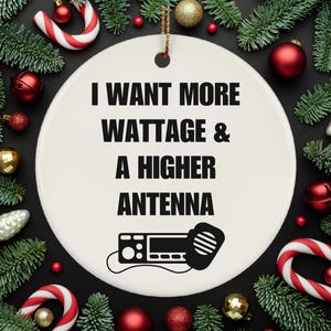 May include: A white ceramic Christmas ornament with the text "I WANT MORE WATTAGE & A HIGHER ANTENNA" in black. A graphic of a radio with a microphone is below the text. The ornament is surrounded by Christmas decorations.