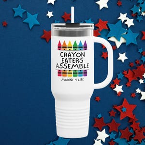 May include: White travel mug with a black lid and handle. The mug features the text "CRAYON EATERS ASSEMBLE" with a colorful crayon design. The phrase "MARINE 4 LIFE" is printed below the text. The background is a dark blue with red, white, and blue star decorations.
