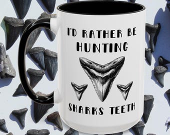 May include: White ceramic mug with a black interior and handle. The mug features the text "I'D RATHER BE HUNTING SHARKS TEETH" with a graphic of a shark tooth. The background is filled with shark teeth.