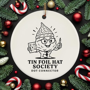 May include: A white ceramic Christmas ornament with a black illustration of a cartoon character wearing a tin foil hat, glasses, and a tie. The text "TIN FOIL HAT SOCIETY DOT CONNECTOR" is below the character. The ornament is surrounded by Christmas decorations.