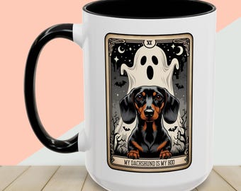 May include: A white ceramic mug with a black handle and interior. The mug features a Halloween-themed design with a dachshund dog, a ghost, and the text "MY DACHSHUND IS MY BOO". The design is framed with a decorative border.