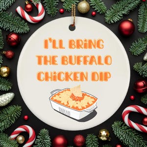 May include: A white ceramic ornament with the text "I'll Bring The Buffalo Chicken Dip" in orange. The ornament features an illustration of a casserole dish filled with dip and tortilla chips, surrounded by Christmas decorations.