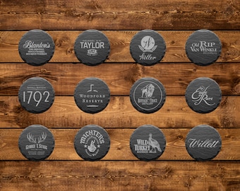 May include: Twelve round black slate coasters with white text. The coasters feature the names of various bourbon brands, including Blanton's, Taylor, Old Rip Van Winkle, 1792, Woodford Reserve, Eagle Rare, Buffalo Trace, E.H. Taylor Jr.,  Michter's, Wild Turkey, and Willett.