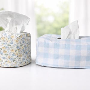 May include: Two tissue box covers. One is cylindrical with a floral pattern in blue, yellow, and white. The other is rectangular with a blue and white gingham pattern. Both have white tissues protruding from the top.