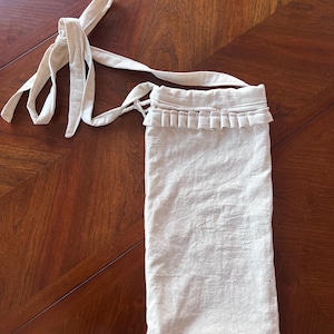 Puede incluir: Una bolsa de lino beige con una correa larga y un detalle de volantes decorativos en la parte superior. La bolsa tiene un cierre de cordón y está hecha de una tela texturizada. La bolsa está diseñada para llevarse al hombro o cruzada.