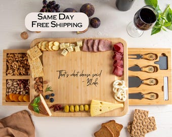 May include: A bamboo cheese board with a slide-out drawer filled with nuts and crackers. The board is laden with cheese, meats, and crackers. A wine bottle and glass are in the background. The board is engraved with the phrase "That's what cheese said - Blake."