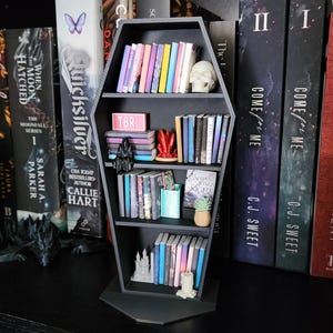 Puede incluir: Una estantería negra en forma de ataúd llena de libros coloridos y objetos decorativos. La estantería tiene varios niveles y está rodeada de otros libros. Se ve una calavera pequeña y la palabra "TBR".