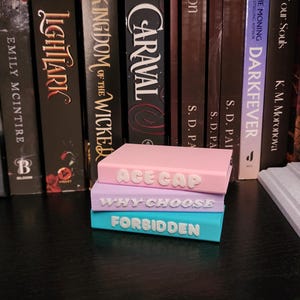 May include: Three stacked, rectangular book-shaped objects in pastel colors: pink, lavender, and teal. Each has white text: "ACE GAP", "WHY CHOOSE", and "FORBIDDEN". The objects are in front of a bookshelf filled with books.
