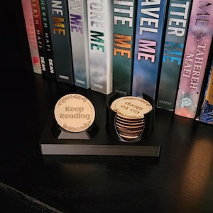 May include: A black coaster holder with wooden coasters. One coaster says "Keep Reading" and another says "Go To Sleep". Books with titles including "Shatter Me" and "Unravel Me" are visible in the background.