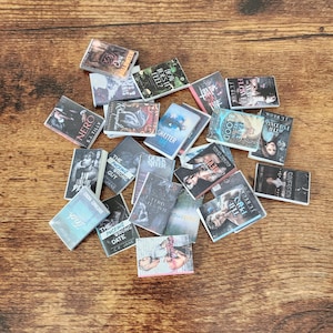 Könnte beinhalten: Eine Ansammlung von Miniaturbüchern mit verschiedenen Titeln und Cover-Designs, die auf einer Holzoberfläche verstreut sind. Titel sind unter anderem "NERO", "SHATTER ME" und "THE WRONG GUY".