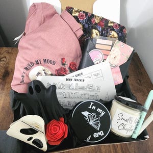 Puede incluir: Caja de regalo con una camiseta rosa con el texto "Don't Mind My Mood", una decoración negra con forma de fantasma, un objeto con forma de calavera, una vela y un rastreador de libros.