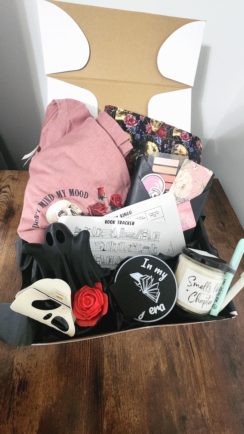 May include: A gift box filled with various items, including a pink t-shirt with the text "Don't Mind My Mood", a black ghost-shaped decoration, a skull-shaped item, a candle, and a book tracker.