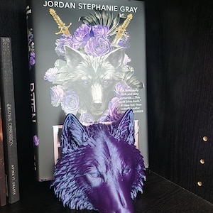 May include: A three-dimensional, metallic purple wolf head sculpture with pointed ears and detailed fur texture. The sculpture is positioned in front of a book titled Jordan Stephanie Gray, which features a wolf illustration and purple roses.