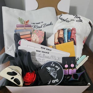 May include: A gift basket filled with book-themed items. Includes a tote bag with the text "I just want to read books", a blanket, a book bingo card, earrings, a ghost-shaped item, a rose, and a patch with the text "In my era".