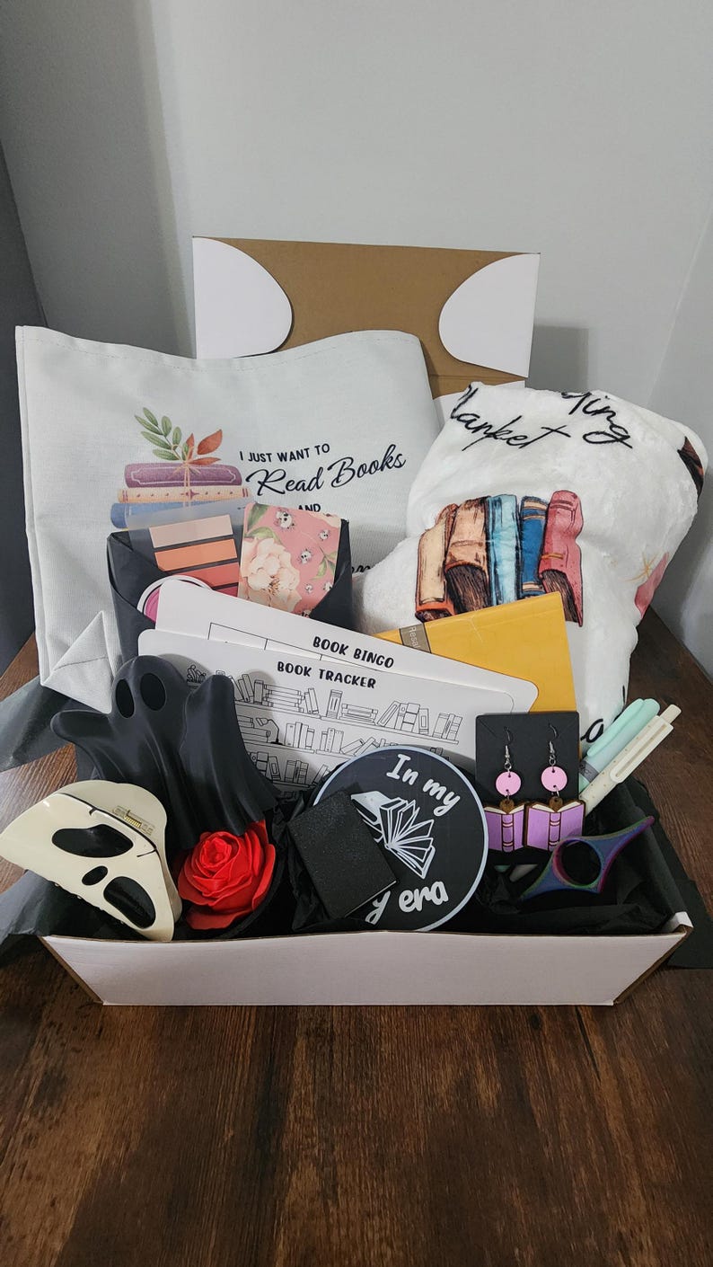 May include: A gift basket filled with book-themed items. Includes a tote bag with the text "I just want to read books", a blanket, a book bingo card, earrings, a ghost-shaped item, a rose, and a patch with the text "In my era".