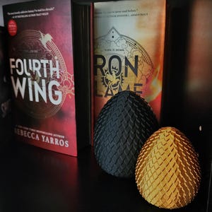 May include: Two dragon egg-shaped bookends, one black and one gold, on a black bookshelf with books behind them. The books are titled "Fourth Wing" and "Iron Flame".