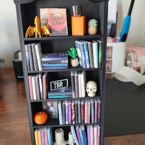 May include: A miniature black bookshelf filled with tiny books, a golden dragon figurine, a small orange pumpkin, a white skull, and a small potted succulent. The books have colorful spines and the text "TBR" is visible on one book.