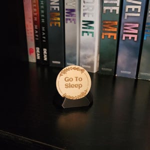 May include: A wooden, circular desk accessory with the text "Go To Sleep" and a floral border. The accessory is displayed on a black surface in front of a row of books.