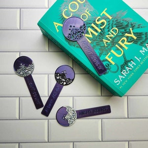 May include: Three purple and white bookmarks with a geometric design and the text "NIGHT COURT" printed on them. The bookmarks are on a white tiled surface. A book with a teal cover and the text "A COURT OF MIST AND FURY" is in the background.