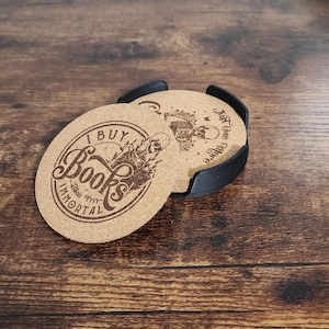 May include: Set of two cork coasters with a black holder. One coaster features the text "I Buy Books Like I'm Immortal" with a skeleton graphic. The other coaster has the text "Just One More Chapter" with a skeleton graphic. The coasters are on a wooden surface.