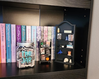 May include: A dark wooden bookshelf with various items. A glass jar labeled "TBR JAR" is filled with slips of paper. Books with colorful spines line the shelf. A small black bookshelf displays miniature items, including a pineapple and a skull.