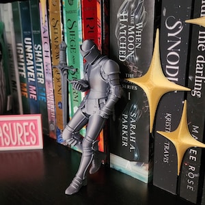 May include: A gray knight figurine in full plate armor stands leaning against a row of books. The knight holds a small object. Two gold-colored, four-pointed star ornaments are positioned near the books. A pink sign with the text ASURES is visible.