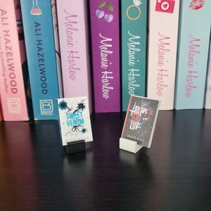 May include: Miniature book replicas, including "Sweet Venom" and "That Girl in Love," are displayed in front of a row of colorful book spines. The spines feature titles and author names, with a pink, blue, and purple color scheme.