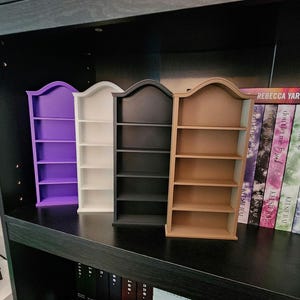 Könnte beinhalten: Vier Miniatur-Bücherregale in Lila, Weiß, Schwarz und Beige. Jedes Regal hat eine geschwungene Oberseite und vier Regale. Die Regale werden auf einem schwarzen Regal mit Büchern im Hintergrund ausgestellt.