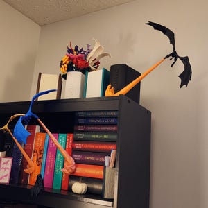 Puede incluir: Una estantería negra con una variedad de libros, incluyendo "El Señor de los Anillos" y "El Hobbit". También hay dos separadores de libros de dragón naranja y azul y un separador de libros de dragón negro en la estantería.