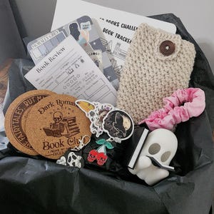 Puede incluir: Una caja de regalo con artículos temáticos de libros. Incluye posavasos de corcho, pegatinas, una funda de teléfono de ganchillo, una goma para el pelo rosa, una figura de fantasma y una hoja de revisión de libros. La caja está forrada con papel de seda negro.