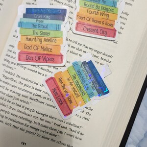 May include: A close-up of a book with several colourful bookmarks. The bookmarks are stacks of books with titles such as "Cruel King", "The Ritual", "The Sinner", "Haunting Adeline", "God of Malice", "Den of Vipers", "From Blood and Ash", "Bite the Woman That Feeds", "Shatter Me", "Bound by Dragons", "Fourth Wing", "Court of Thorns and Roses", "Crescent City", "The Amendment", "Twenty Years Later", "The Girl Who Was Taken", "Watching You", "The Housemaid", "In An Instant", and "Then She Was Gone".