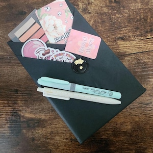 May include: A black envelope sealed with a gold-flecked wax seal, containing stickers, bookmarks, and two pens. The stickers feature floral and ghost designs. One pen is light blue, and the other is white with small dots. The envelope has the word "Reading" printed on it.