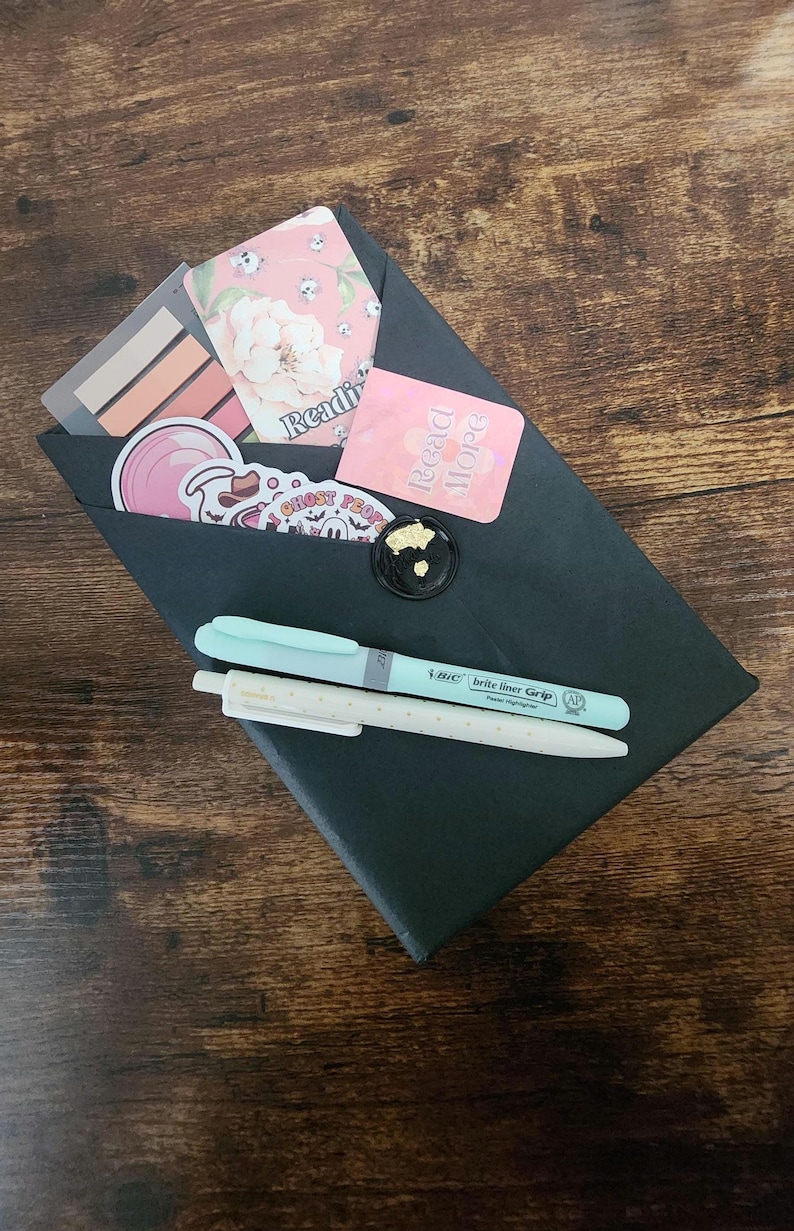 May include: A black envelope sealed with a gold-flecked wax seal, containing stickers, bookmarks, and two pens. The stickers feature floral and ghost designs. One pen is light blue, and the other is white with small dots. The envelope has the word "Reading" printed on it.