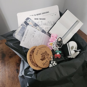 May include: A collection of book-themed items, including a book tracker, book review sheets, coasters with "Dark Romance Book Club" and "Tales But Made Fairytales" text, stickers, and a ghost figurine reading a book. The items are arranged on a dark surface.