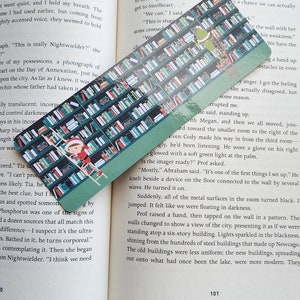 May include: A green bookmark featuring a bookshelf design with a girl in a red and white Santa suit climbing a ladder. The bookmark is placed on top of an open book with text about a city.