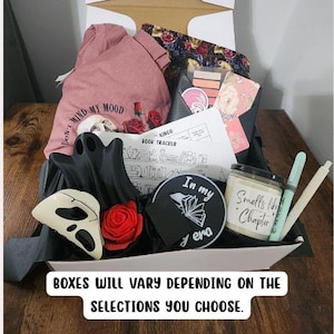 May include: A gift box filled with various items, including a pink t-shirt with a skull and rose graphic, a black ghost-shaped item, a candle, and a book tracker. The box also contains a black and white round item with the text "In my era".