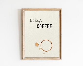 Puede incluir: Una impresión enmarcada con el texto "but first, COFFEE" y una mancha de café en forma de anillo. La impresión está sobre un fondo beige claro y el marco es de madera marrón claro.