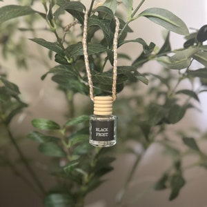 May include: A glass car air freshener with a wooden top and a black label that says "Black Frost". The air freshener is hanging from a string with gold accents.