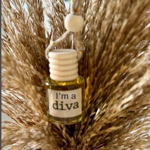 May include: A small, clear glass bottle with a wooden cap and the text "I'm a diva" on a label. The bottle is filled with a golden liquid and is suspended in front of a backdrop of dried, feathery brown grasses.