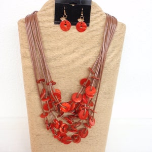 May include: A multi-strand necklace with red wooden disc beads and matching earrings. The necklace is displayed on a brown, woven stand.