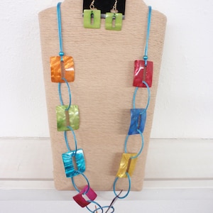 May include: A turquoise blue necklace with colorful rectangular beads. The necklace is made of a turquoise blue cord and has a variety of colors including orange, yellow, green, and blue. The necklace is displayed on a beige stand with a pair of green earrings.