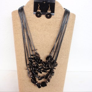 May include: A multi-strand necklace with black round beads and gold accents. The necklace is displayed on a brown stand with a pair of matching earrings.