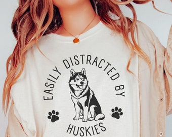 May include: Cream-colored t-shirt with the text "EASILY DISTRACTED BY HUSKIES" in a circular design, featuring a black and white husky illustration and paw prints. The shirt is worn with a tan corduroy jacket.