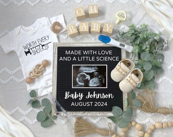 May include: A black letter board with the text "Made with love and a little science" and a sonogram image. Below the text is "Baby Johnson August 2024". There are baby shoes, a pregnancy test, a baby onesie, and other baby items surrounding the letter board.