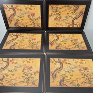 May include: A set of six rectangular placemats with a black border. Each placemat features a yellow background with a floral and tree design in red, white, and brown. The placemats are neatly stacked.